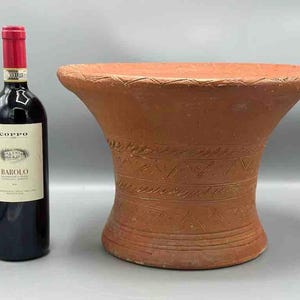 May include: A terracotta pedestal with a decorative pattern, featuring a wide, flat top and a flared base. The pedestal is a reddish-brown color and has a rustic, handmade appearance.