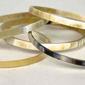 May include: A set of five horn bangles in various shades of cream and gray. The bangles are stacked on top of each other.