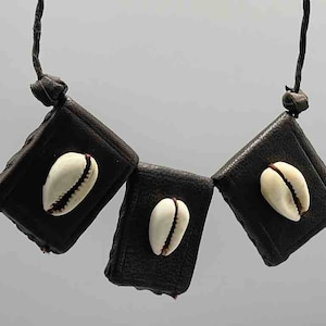 Puede incluir: Un collar con tres colgantes cuadrados oscuros, cada uno adornado con una concha de cauri. Los colgantes están unidos por un cordón oscuro. Las conchas de cauri son blancas con un borde marrón.