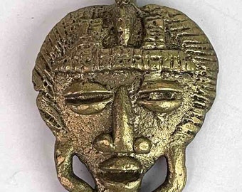 Puede incluir: Un colgante de metal dorado que presenta un rostro humano estilizado con una superficie texturizada. El colgante tiene un bucle en la parte superior para colgar.
