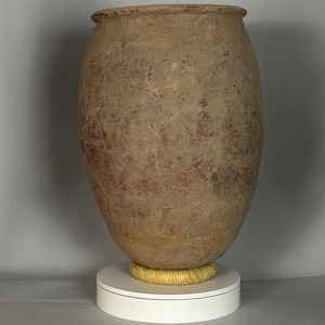 May include: A large, antique-style, beige-colored ceramic urn with a wide mouth and rounded body. The urn has reddish-brown markings and sits on a white pedestal with a gold-colored woven ring. The urn is likely for decorative purposes.