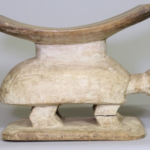 May include: A wooden stool carved in the shape of a turtle. The seat is a wide, flat, curved piece of wood that sits atop the turtle's back. The turtle has four legs and a long neck with a head that is slightly open.