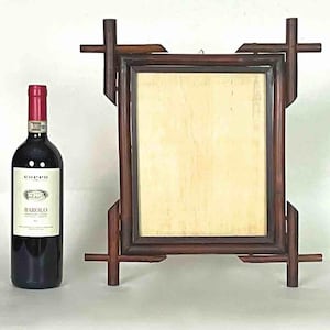 Puede incluir: Un marco de fotos de madera marrón oscuro con un interior de color claro y una botella de vino tinto con una etiqueta blanca. El marco tiene un diseño único con detalles en forma de cruz en los lados. La botella de vino tiene una tapa roja y una etiqueta que dice "Barolo".