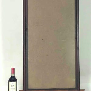Puede incluir: Un espejo alto y rectangular con marco de madera oscura y un panel central marrón claro. Una botella de vino tinto se encuentra a la izquierda del espejo. El espejo tiene soportes horizontales en la parte superior e inferior.