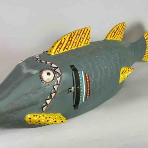 Puede incluir: Una escultura de pescado de madera pintada a mano con un cuerpo gris, acentos amarillos y rojos y una cara detallada. El pez tiene una boca grande con dientes afilados y un solo ojo.