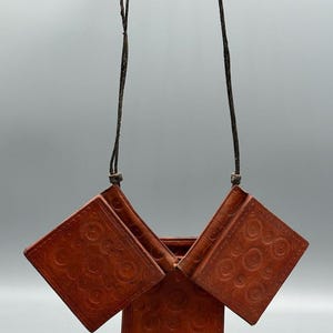May include: A hanging, open, brown leather wallet with a decorative embossed floral pattern. The wallet is suspended by a thin, dark brown cord. The wallet's design includes multiple square compartments.