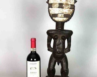 May include: A dark wood sculpture of a human figure with horns and a white face mask. The sculpture is standing on a base with intricate carvings. A bottle of wine is in the foreground.