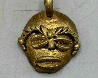 Puede incluir: Un colgante dorado con un rostro estilizado con un ojo cerrado y un bucle para colgar.