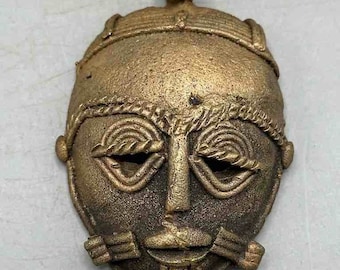 Puede incluir: Un colgante de máscara de color bronce con un lazo en la parte superior para colgar. La máscara presenta rasgos faciales estilizados, incluyendo ojos cerrados, una nariz y una boca. La máscara tiene una superficie texturizada y parece ser un artículo antiguo o vintage.