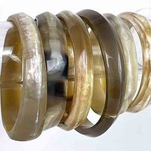 Op de afbeelding: Een set van zeven bruine en crèmekleurige hoornarmbanden. De armbanden zijn op elkaar gestapeld en worden getoond op een transparante acrylstandaard.