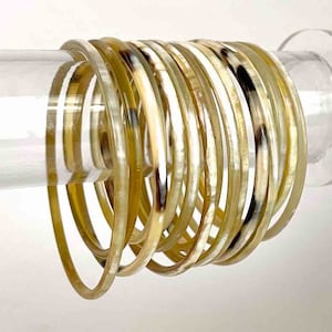 Puede incluir: Un conjunto de 15 brazaletes de cuerno de color marrón claro con detalles en marrón oscuro. Los brazaletes están apilados uno encima del otro y se muestran en un soporte de acrílico transparente.