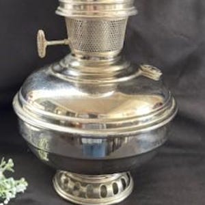 May include: A vintage-style oil lamp with a clear glass chimney and a silver-colored metal base. A tall, olive-green candle is inside the glass. The lamp has a round base and a small handle on the side.