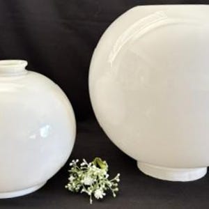 May include: Two white, spherical glass light fixtures. One is smaller with a neck, and the other is larger with a flat base. A small sprig of white flowers sits between them. The fixtures have a glossy finish.