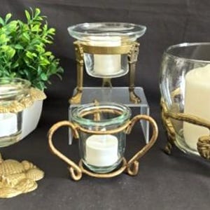 May include: A collection of four glass candle holders with gold-toned metal bases. One base features a seashell design, one has a pedestal with ornate legs, one includes two curved handles, and the largest features a grape vine motif with a white candle.