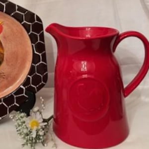 Puede incluir: Una jarra de cerámica roja y un plato decorativo con un diseño de gallo. El plato tiene un borde hexagonal marrón con un patrón de panal blanco. El gallo es amarillo, marrón y negro.