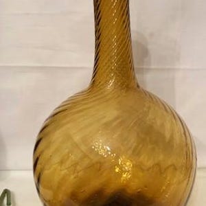 May include: Amber-colored glass bottle with a round body and a long, spiraled neck. The bottle has a vintage aesthetic, with a dark base and a textured surface. A decorative piece for home decor.