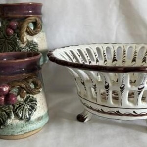 May include: Two ceramic mugs and a decorative bowl. The mugs have a textured handle and are decorated with grape vines and purple accents. The bowl is white with a brown rim and decorative cutouts, resting on three small feet.
