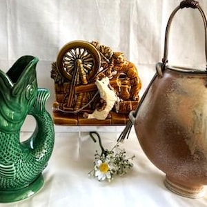 May include: A collection of decorative ceramic items. A green fish-shaped pitcher, a brown ceramic relief sculpture, and a tan and brown ceramic vase with a handle. The items are arranged on a white surface.