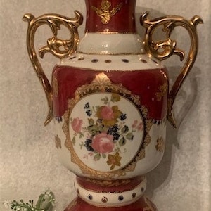 以下が含まれることがあります： 赤と金の磁器の花瓶で、花柄と金のアクセントが施されています。花瓶にはハンドルが2つと幅広のベースが付いています。