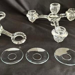 May include: Clear glass candle holders with three arms and a central cup, along with several clear glass drip catchers. The candle holders have a decorative design and are suitable for taper candles. The items are displayed on a dark surface.