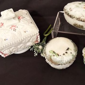 May include: An assortment of antique white ceramic decorative boxes. The boxes feature floral and embossed designs, with pink and green accents. One box is square with a lid, and others are oval and rounded.
