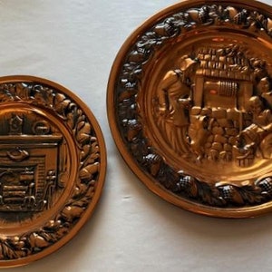 May include: Two copper wall plates with embossed designs. The larger plate features a scene of people working at a well, while the smaller plate depicts a fireplace with a spinning wheel.