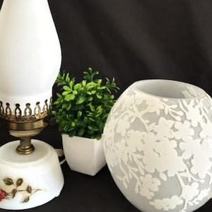 May include: A tall electric lamp with a white frosted glass chimney, a brass base, and a rose decal on the bottom, alongside a small green plant in a white pot and a spherical glass lamp shade featuring a white floral pattern on frosted glass.