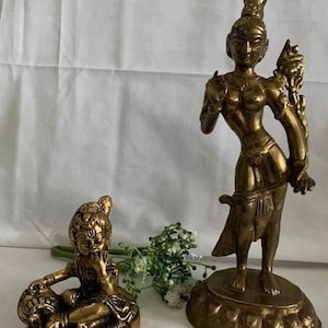 Puede incluir: Dos estatuas de metal dorado de deidades hindúes. La estatua más grande representa a una diosa con una corona y joyas elaboradas. La estatua más pequeña representa a un dios sentado con las manos juntas frente a él.