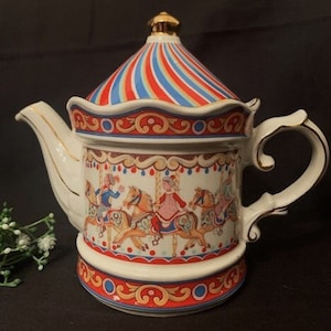 Op de afbeelding: Een witte porseleinen theepot met een kleurrijk carrouselontwerp. De theepot heeft een gouden deksel en handvat en heeft een rood, blauw en geel gestreept baldakijn.