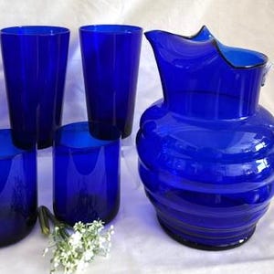 May include: A set of cobalt blue glass drinkware, including a pitcher and glasses. The pitcher has a clear glass handle and a ridged body. There are four tall glasses and two shorter glasses. The set is a vibrant shade of blue.