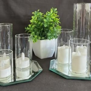 Op de afbeelding: Een set van heldere glazen cilindervazen van verschillende hoogtes, geplaatst op twee achthoekige spiegelplateaus. Elke vaas bevat een wit LED-theelichtje. Een kleine kunstmatige groene plant in een witte vierkante pot staat tussen de twee plateaus.