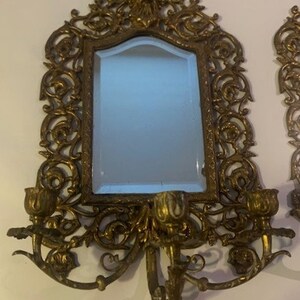 Puede incluir: Un par de apliques de pared de color dorado ornamentadas con un espejo en el centro. Cada aplique tiene un dise&ntilde;o floral decorativo y presenta dos portavelas.