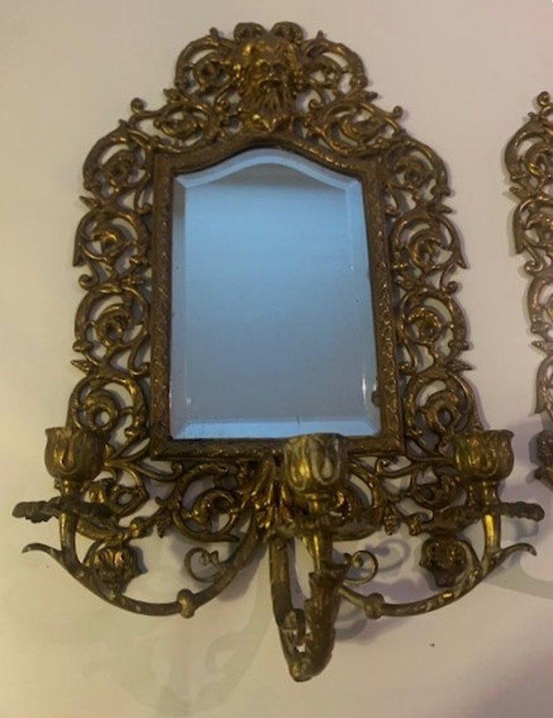 Puede incluir: Un par de apliques de pared de color dorado ornamentadas con un espejo en el centro. Cada aplique tiene un dise&ntilde;o floral decorativo y presenta dos portavelas.