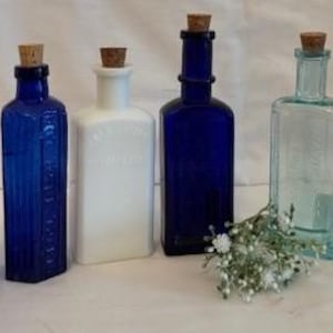 May include: A selection of vintage glass bottles with cork stoppers. The bottles are in shades of blue, clear, and white. A small bunch of white flowers is placed in front of the bottles. The bottles are of various shapes.