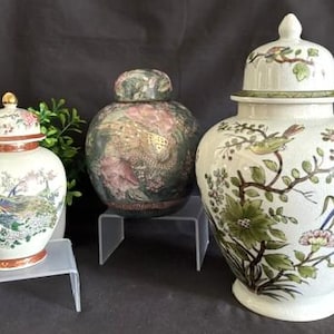 May include: Three ceramic ginger jars with lids. The left jar is white with a peacock and floral design. The centre jar is dark green with a floral pattern. The right jar is white with a bird and floral motif. Each jar sits on a clear stand.