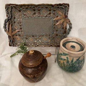 May include: A set of three ceramic pottery pieces: a brown tray with a woven design, a brown jar with a lid, and a green and white jar with a lid.