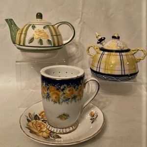 Könnte beinhalten: Ein Set aus drei Teetassen und Untertassen mit Blumendesigns. Die Teekanne ist grün und weiß mit gelben Rosen, die Zuckerdose ist gelb und weiß mit einem karierten Muster und die Teetasse ist weiß mit blauen und gelben Rosen.