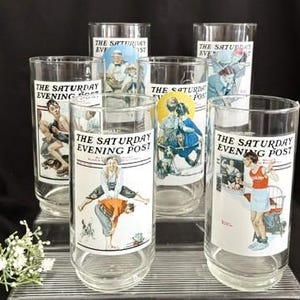 May include: Set of six clear glass tumblers featuring vintage illustrations from The Saturday Evening Post. Each glass displays a different colorful image with the publication's logo. The glasses are arranged on a clear, ridged surface against a dark background.