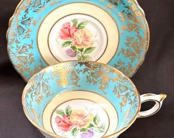 May include: A vintage teacup and saucer set. The set features a light blue exterior with gold floral accents. The interior is cream-colored with a floral design in the center. The cup has a gold handle and base, and the saucer has a scalloped edge.