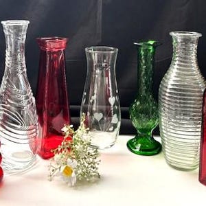 May include: Collection of decorative glass vases in various shapes and colors. Includes red, clear, and green vases. Some vases have textured designs, while others are smooth. A small bouquet of white flowers is in front of the vases.