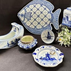 Puede incluir: Colección de objetos decorativos de cerámica azul y blanca. Incluye una fuente con forma de pez, un plato pequeño con un velero, un cenicero pequeño, un tarro pequeño con tapa y una jardinera curvada. Estilo Delftware.