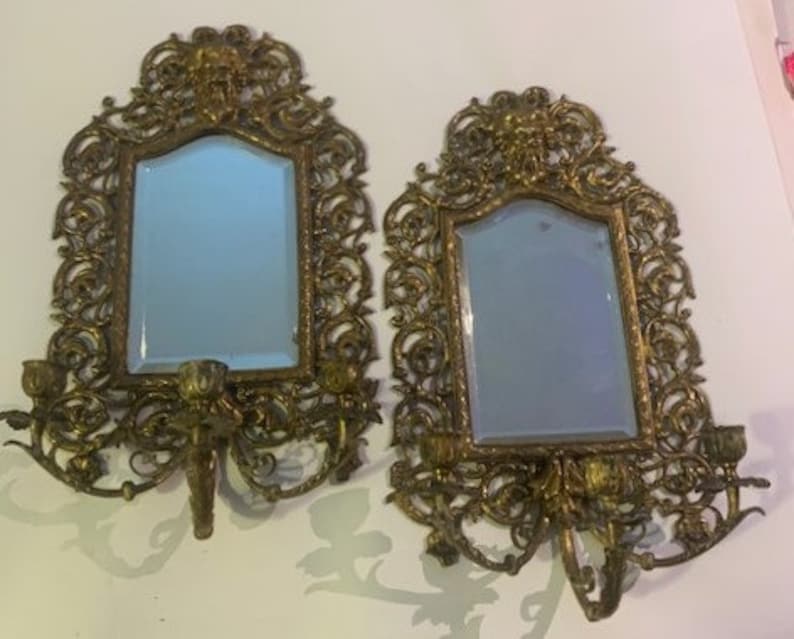 Puede incluir: Un par de espejos de pared ornamentados de color dorado con candelabros. Cada espejo tiene un marco decorativo con detalles intrincados y un espejo de vidrio biselado.