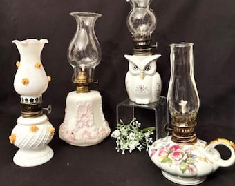 Könnte beinhalten: Eine Sammlung antiker Öllampen. Die Lampen haben weiße Porzellansockel mit floralen und geprägten Details, klare Glaszylinder und Messingakzente. Eine Lampe ist eulenartig geformt. Die Lampen sind auf dunklem Untergrund angeordnet.
