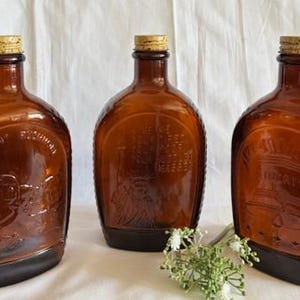 May include: Three amber glass bottles with cork stoppers. Each bottle has a different embossed design on the front. The bottles are on a white surface.