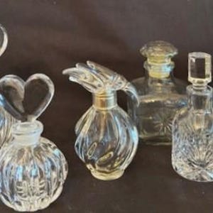 May include: Six clear glass perfume bottles with various shapes and stoppers. The bottles are on a dark surface.