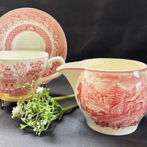 May include: A vintage tea set with a red and white pattern. The set includes a teacup, saucer, and creamer. The teacup and creamer feature a scenic design. The set is displayed on a dark surface.