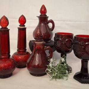 Puede incluir: Conjunto de cinco botellas y copas de vidrio rojo rubí. Las botellas tienen un patrón de diamante y las copas tienen un patrón floral. Las botellas tienen tapones con una parte superior puntiaguda.