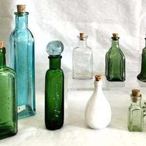 Peut inclure: Collection de bouteilles en verre vintage de différentes formes et tailles, avec des bouchons en liège. Les bouteilles sont de couleurs vertes, bleues et transparentes, disposées sur une surface blanche. Idéal pour la décoration intérieure.