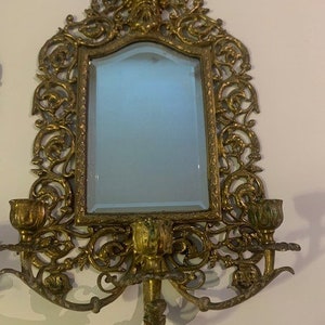 Puede incluir: Espejo de pared de metal dorado adornado con un marco de volutas decorativas y dos candelabros. El espejo tiene un borde biselado y una superficie de vidrio transparente.
