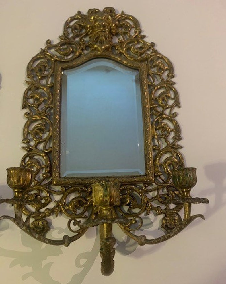 Puede incluir: Espejo de pared de metal dorado adornado con un marco de volutas decorativas y dos candelabros. El espejo tiene un borde biselado y una superficie de vidrio transparente.